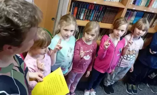 Uczniowie klas pierwszych stoją na tle regałów z książkami, mają uniesione palce do przysięgi. 