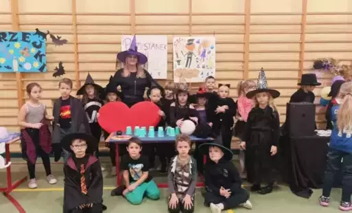 Klasa 1 z wychowawcą przy stoisku z wróżbami, uczniowie ubrani w ciemne szaty i z kapeluszami. 