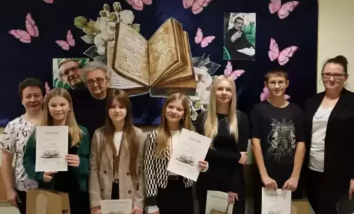 Zwycięzcy i wyróżnieni uczniowie pozują z nagrodami na tle dekoracji - rozłożonej książki, motyli i fotografii Jana Twardowskiego 