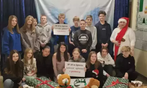 grupa uczniów pozuje z opiekunką koła panią Renatą Gieragą, w tle niebieska tkanina. Uczniowie trzymają prezenty i kartki. 