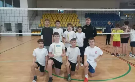 Ósemka uczniów w dwóch rzędach pozuje na tle siatki od siatkówki, trzymają dyplom, obok stoi nauczyciel wychowania fizycznego