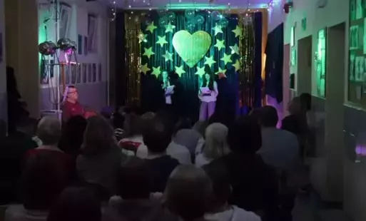 W tle w dekoracja z sercem, dwoje uczniów prowadzących na scenie, rodzice na widowni.