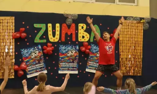 Na scenie prowadzący zajęcia, w tle kolorowy napis zumba