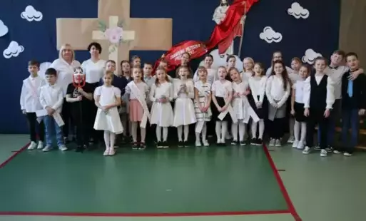 Na tle dekoracji stoją ubrane na biało dzieci z klasy pierwszej wraz z nauczycielkami przygotowującymi spektakl. 