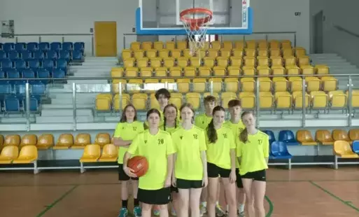 Drużna chłopców i dziewczynek w żółtych strojach sportowych, z piłką do koszykówki w ręku, stoją na tle trybun.