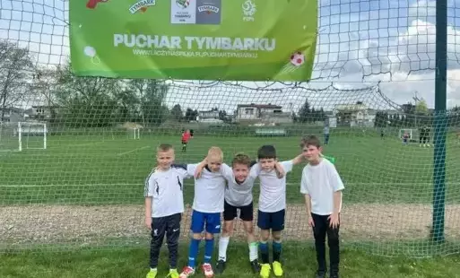 Pięciu chłopców w sportowych strojach stoją na tle murawy piłkarskiej.