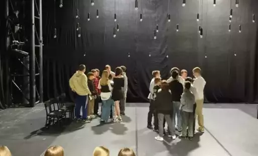 Dwie grupki uczniów na scenie, pod sufitem zawieszone żarówki. 