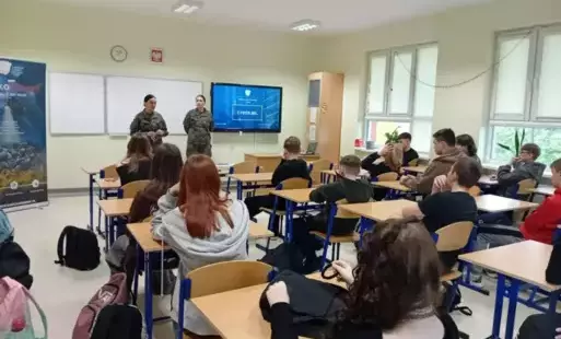 Uczniowie siedzą w ławkach, przy tablicy dwie panie w mundurze Wojska Polskiego.