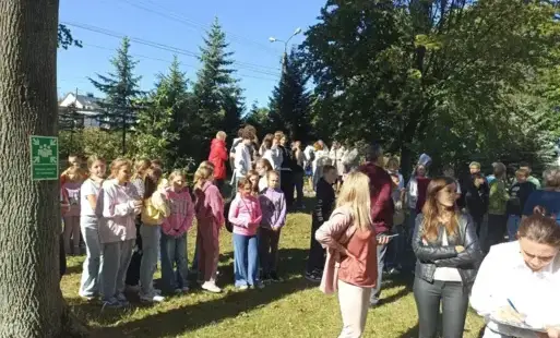 uczniowie i nauczyciele na zewnątrz stoją przed znakiem "miejsce zbiórki ewakuacyjnej" 