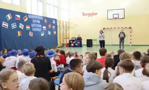 Dzieci siedzące na sali gimnastycznej, dwie nauczycielki prowadzące uroczystość, z boku napis i flagi krajów europejskich 