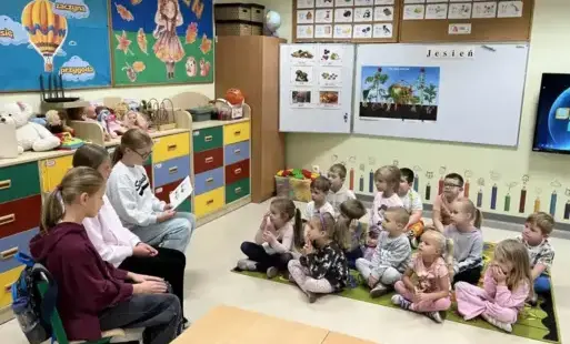 Trzy uczennice siedzą z boku i czytają, przedszkolaki siedzą na dywanie i słuchają 