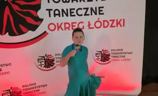 Uczennica w sukience pozuje z medalem na tle ścianki z napisem o konkursie 