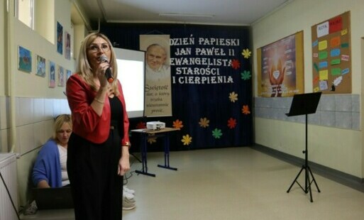 Na piewszym tle pani dyrektor z mikrofonem, z tyłu dekoracja i baner z wizerunkiem papieża.