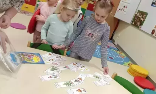 Kilkoro dzieci stoi przy stoliczku i pochyla się nad obrazkami leżącymi na blacie. 